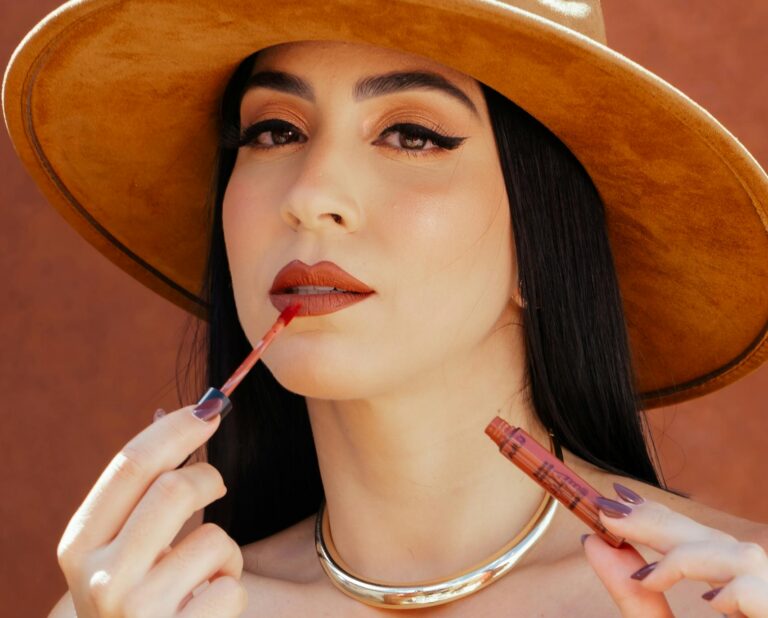 Elegant woman wearing chic wide-brim hat applying lipstick with confidence and style.